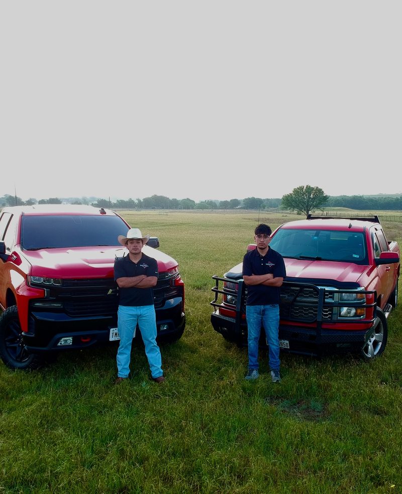 Texas Junk Boyz crew in branded polos standing between two red Chevy trucks in a Texas field